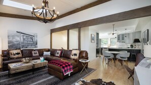 A spacious living room with a brown leather sofa, a wooden coffee table, and a chandelier. There is a dining area with a table and chairs, and a kitchen in the background.