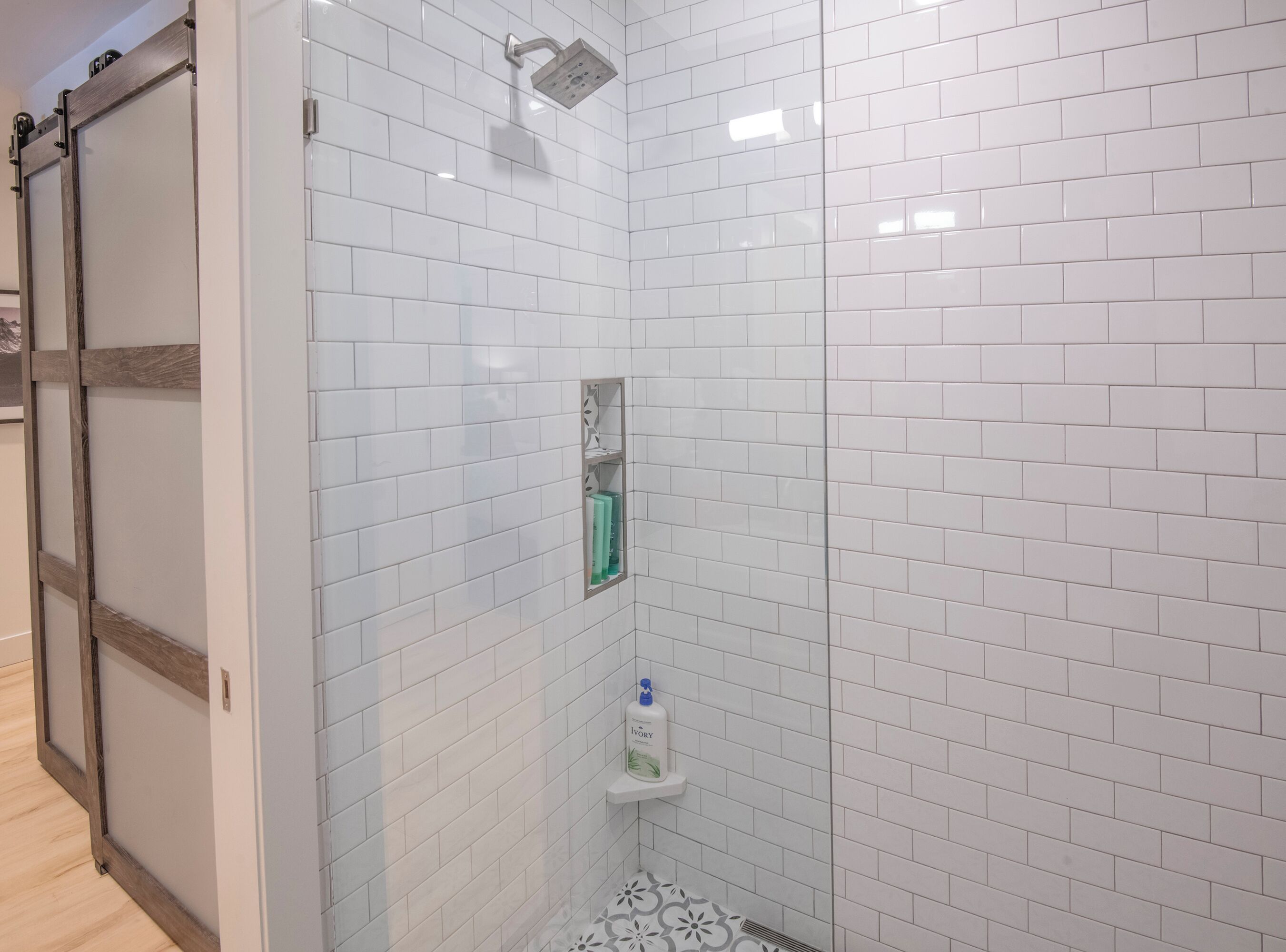 A bathroom with white subway tiles, a shower with a handheld showerhead, and a shelf with toiletries.