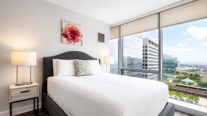 Et moderne hotelværelse med en stor seng, to sengeborde med lamper, udsigt over byen og et blomsterbillede på væggen.