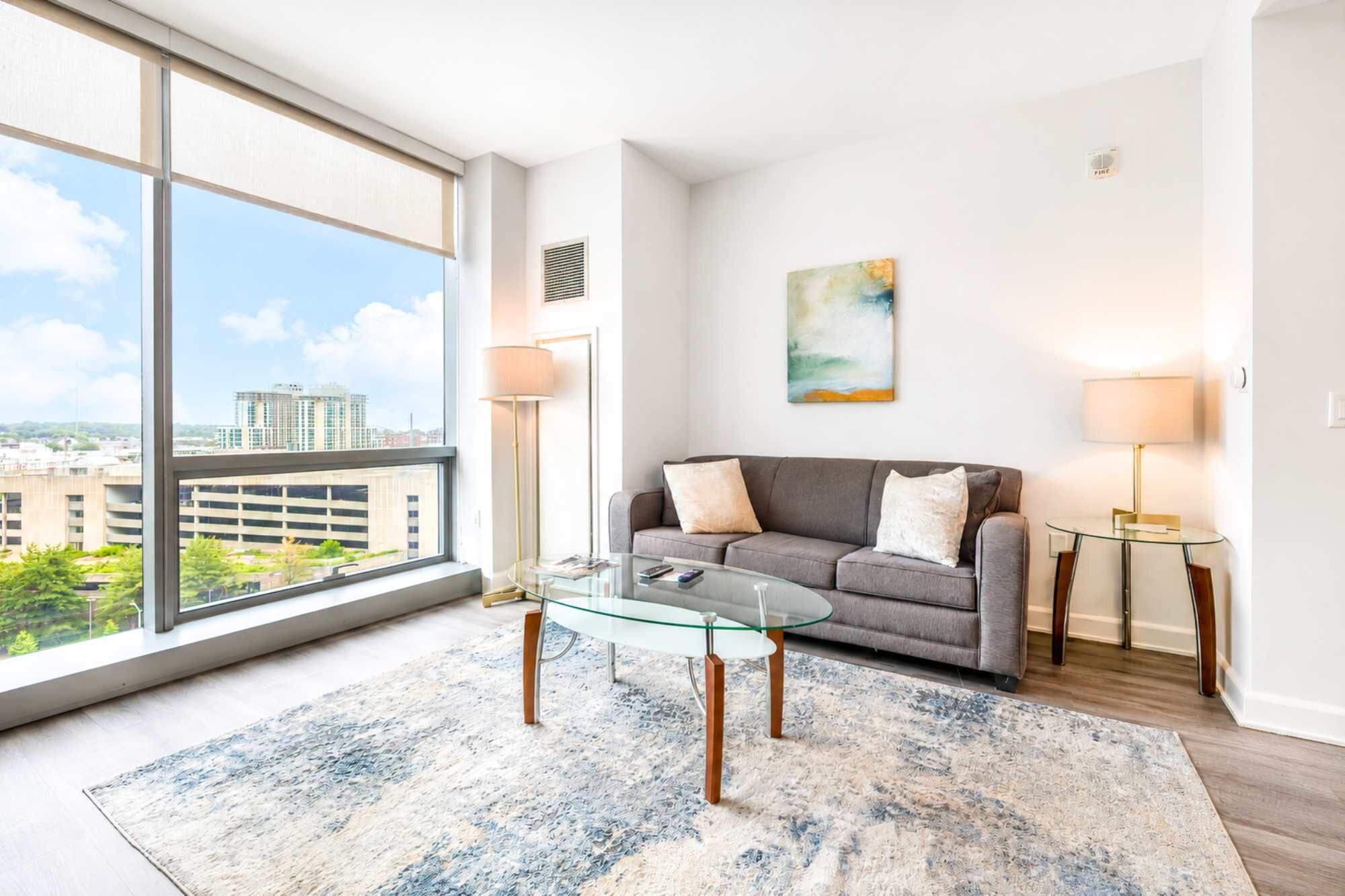 A modern living room with a glass coffee table, grey sofa, and a large window offering a city view.