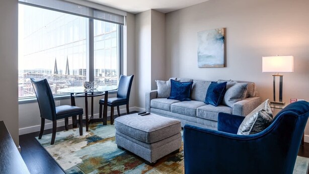 A modern living room with a sofa, chairs, a coffee table, and a view of a cityscape through large windows. - Private Downtown Seaport Apt with Pool (Boston MA)