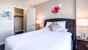 A hotel room with a bed, a nightstand, a bedside lamp, a door, and a wall with a painting. - Private Downtown Seaport Apt with Pool (Boston MA)