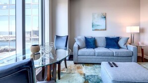 A modern living room with a grey sofa, a glass table, and a city view. - Private Downtown Seaport Apt with Pool (Boston MA)