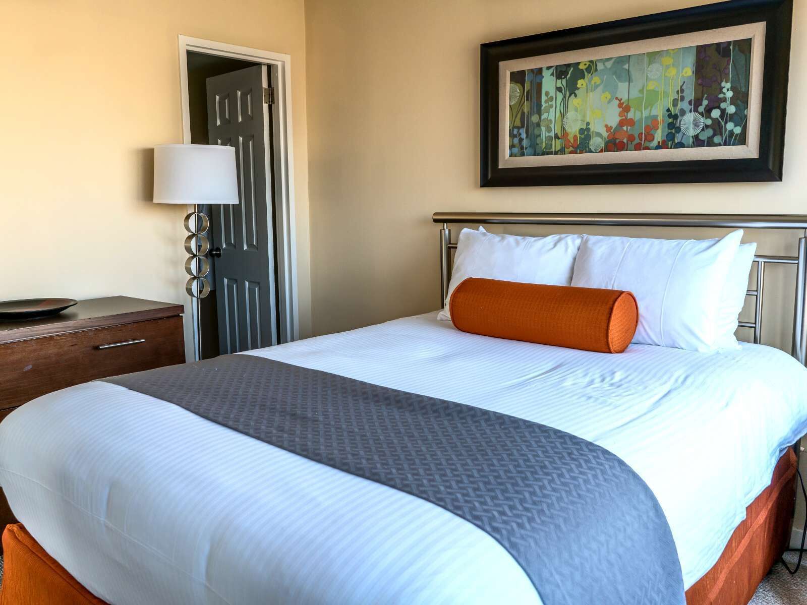 A neatly made bed with white and orange bedding, a metal headboard, and a framed abstract painting above it.