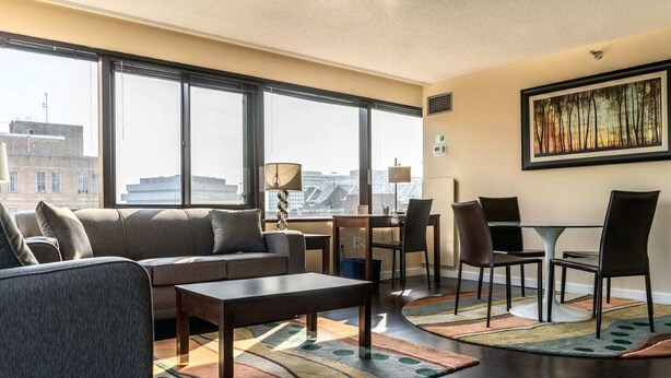 Habitación de hotel moderna con sofá, mesa de centro, mesa de comedor, sillas y ventanas amplias con vistas a la ciudad.