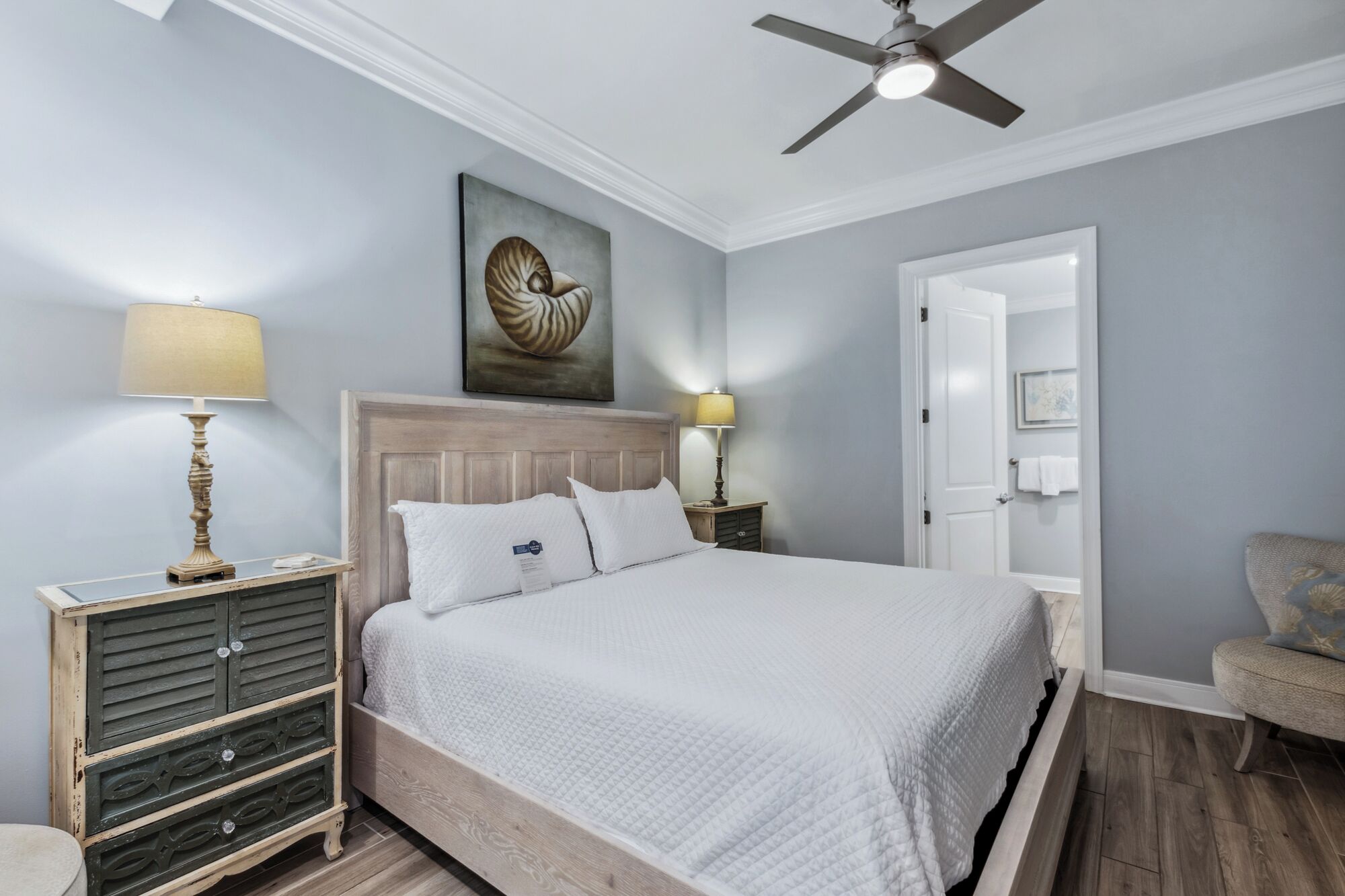 Une chambre à coucher bien rangée, avec un lit, des lampes de chevet, un ventilateur de plafond et une porte donnant sur une autre pièce.