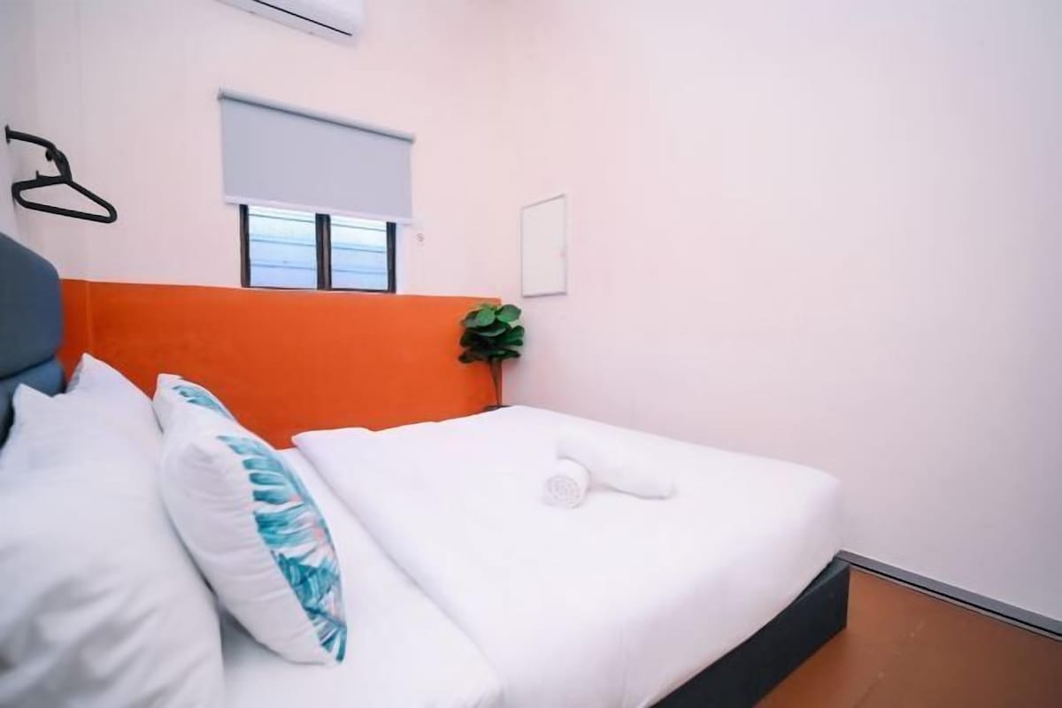 Une chambre d’hôtel avec un lit, du linge de lit blanc, un oreiller bleu et blanc, une serviette blanche, une petite plante verte, une fenêtre avec un store et un porte-manteau fixé au mur.