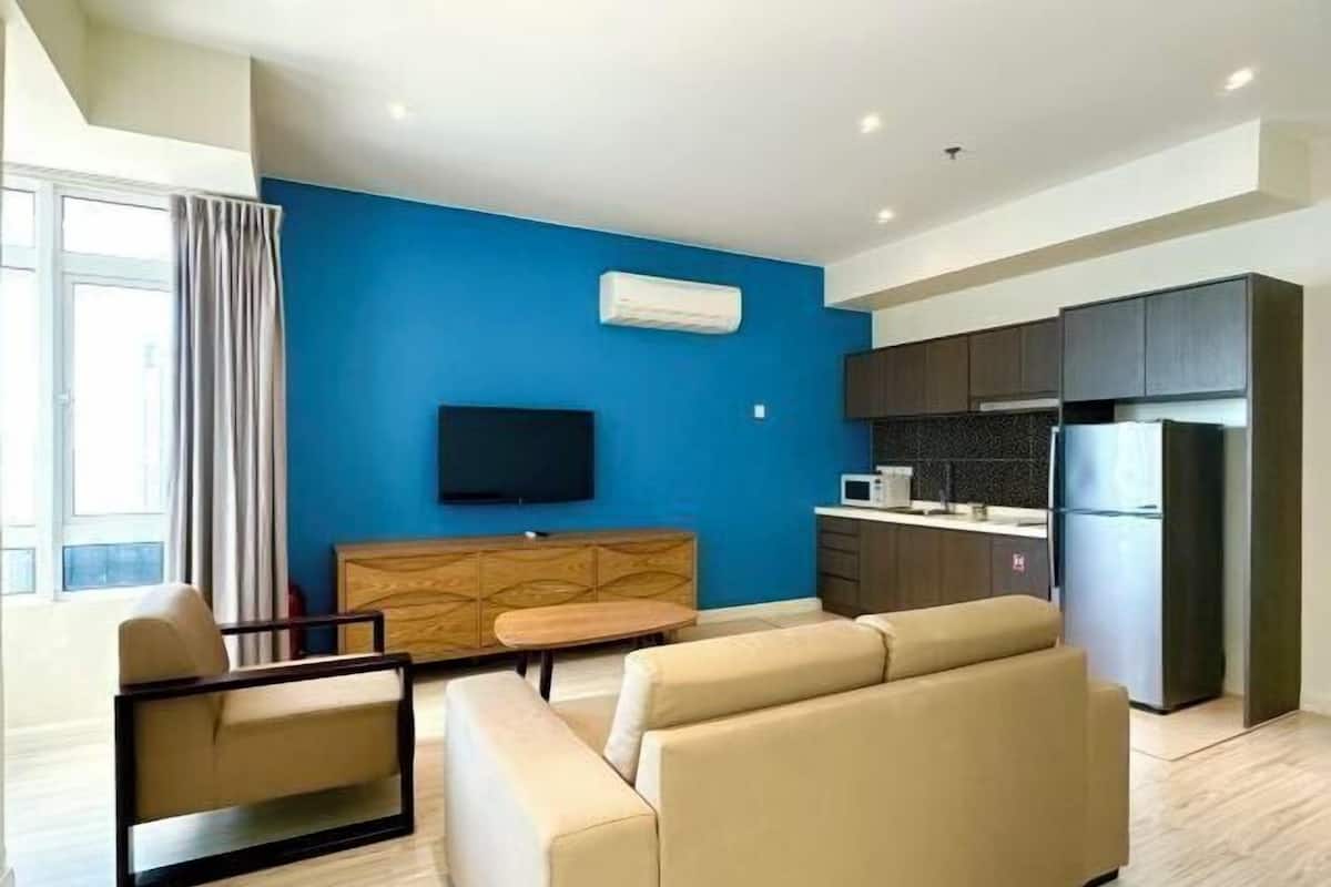 Un soggiorno moderno con una parete azzurra a contrasto, una TV a schermo piatto, un divano, un tavolino da caffè e un angolo cottura con mobili scuri e un frigorifero.