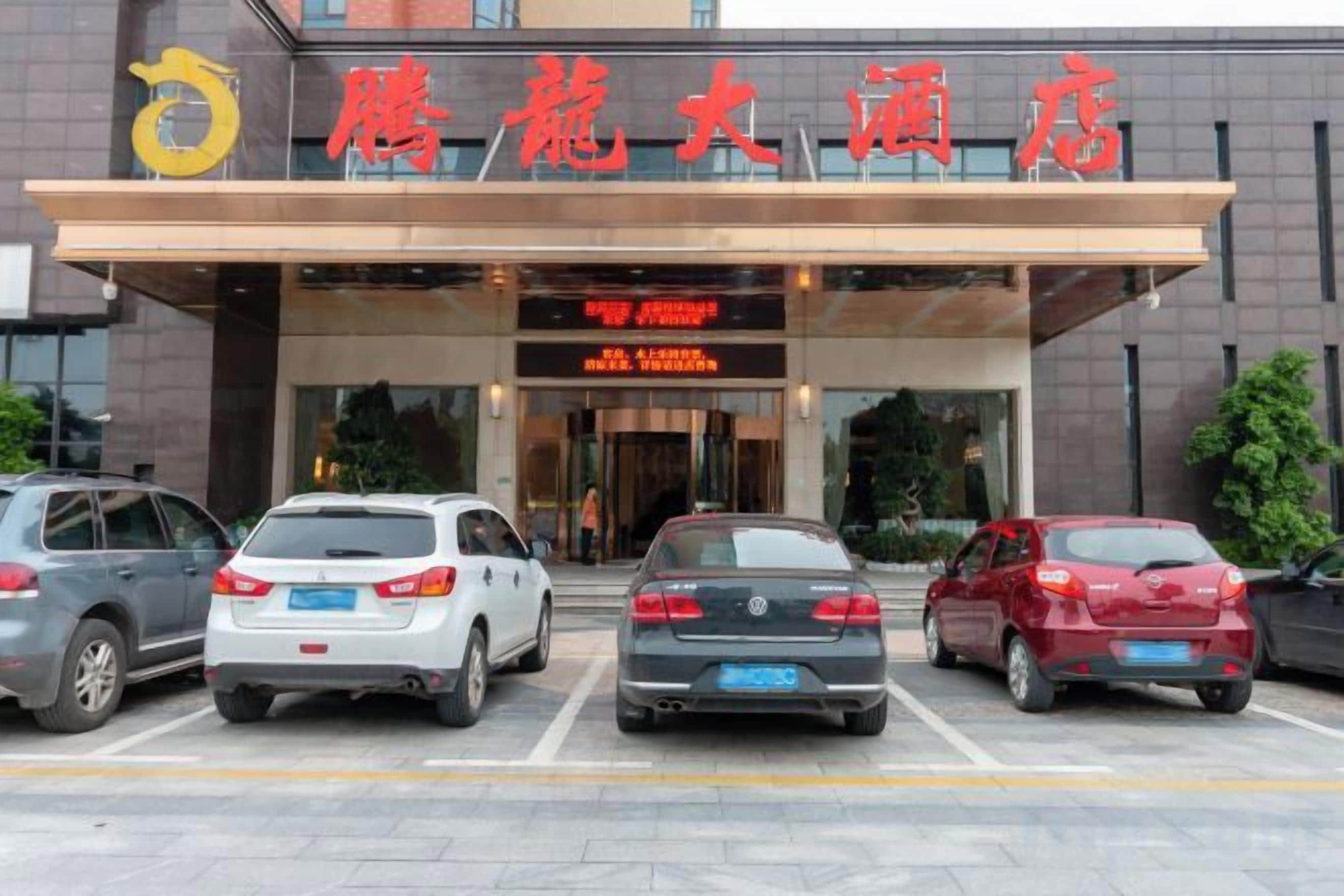 A hotel entrance with a clear sign, a parking lot with several cars, and a modern building facade.