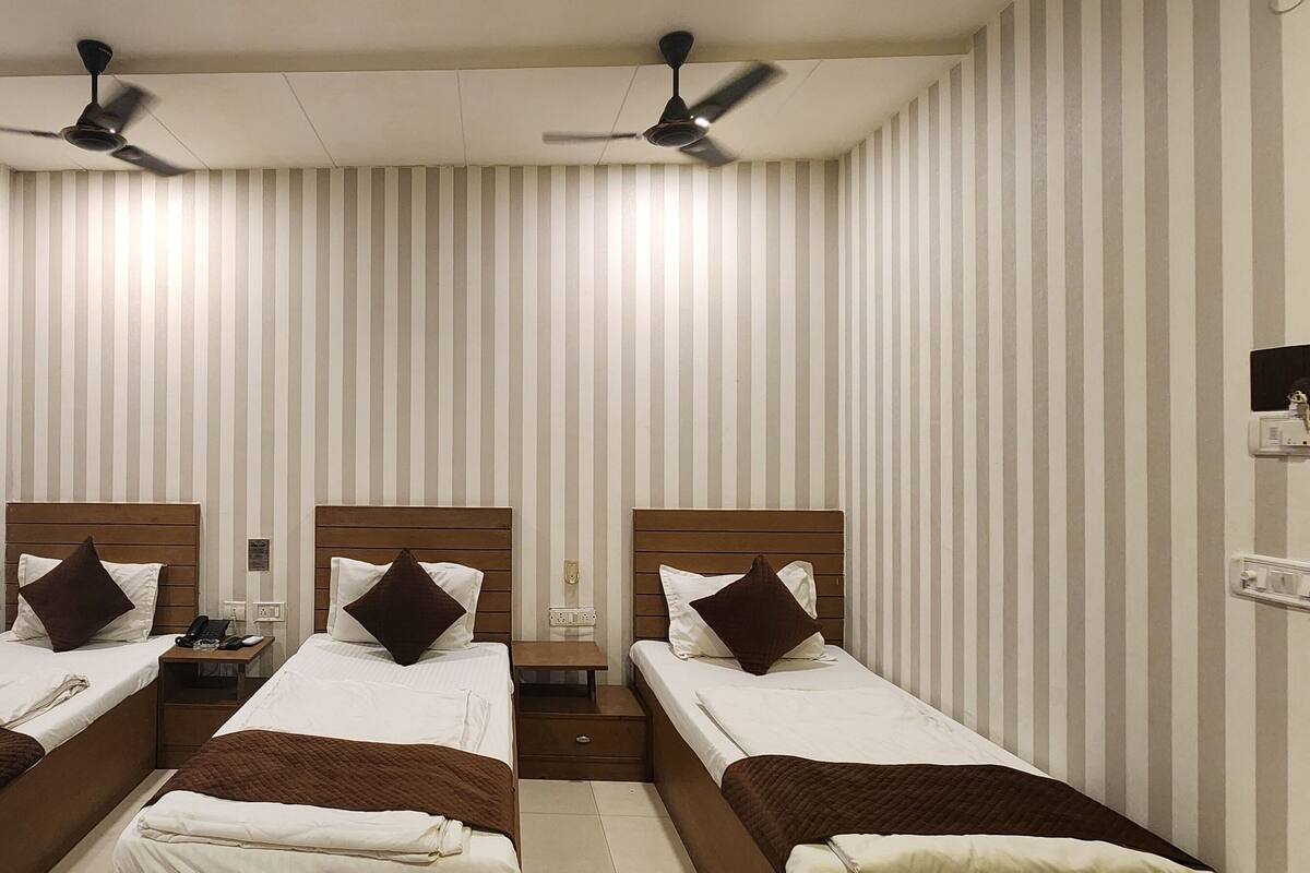 A hotel room with three single beds, each with a wooden headboard, white bedding, and brown throw blankets. There are ceiling fans above each bed, and a clock on the wall.