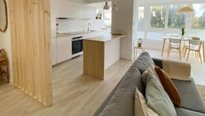Une cuisine moderne avec des meubles blancs et un îlot central en bois. Le salon est agrémenté d’un canapé gris avec des coussins, et une table à manger avec des chaises se trouve à l’arrière-plan.