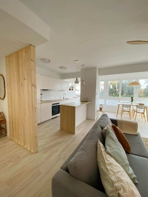 Une cuisine moderne avec des meubles blancs et un îlot central en bois. Le salon est agrémenté d’un canapé gris avec des coussins, et une table à manger avec des chaises se trouve à l’arrière-plan.