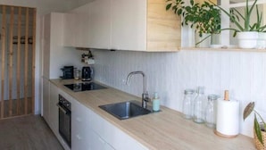 Une cuisine moderne avec des meubles blancs, un plan de travail en bois et un évier encastré. On aperçoit des plantes en pot sur une étagère et une porte en bois au fond.