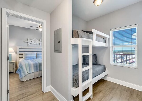 A room with bunk beds, a bed with striped bedding, a small desk with a lamp, a starfish decoration, and a window with a view of the beach.