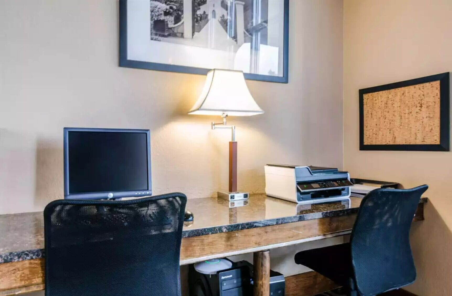 Una habitación de hotel con un escritorio, dos sillas, una lámpara, una impresora, un monitor y cuadros en la pared.