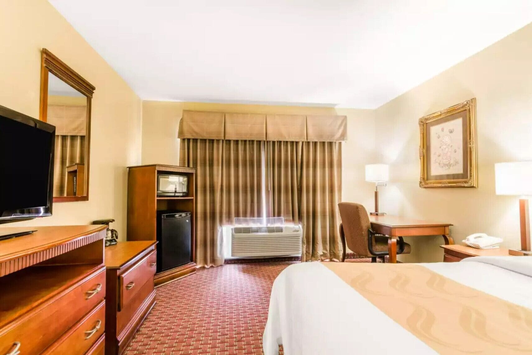 A hotel room with a bed, a desk, a television, a minibar, and a window with curtains.