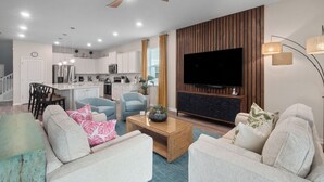 A modern living room with a flat-screen TV, a ceiling fan, and a kitchen in the background.