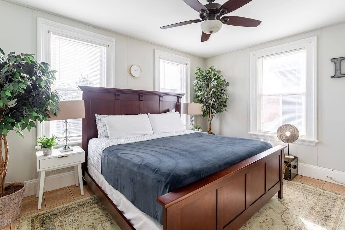 Une chambre à coucher avec un lit en bois, des tables de chevet blanches, un ventilateur de plafond et de grandes fenêtres.