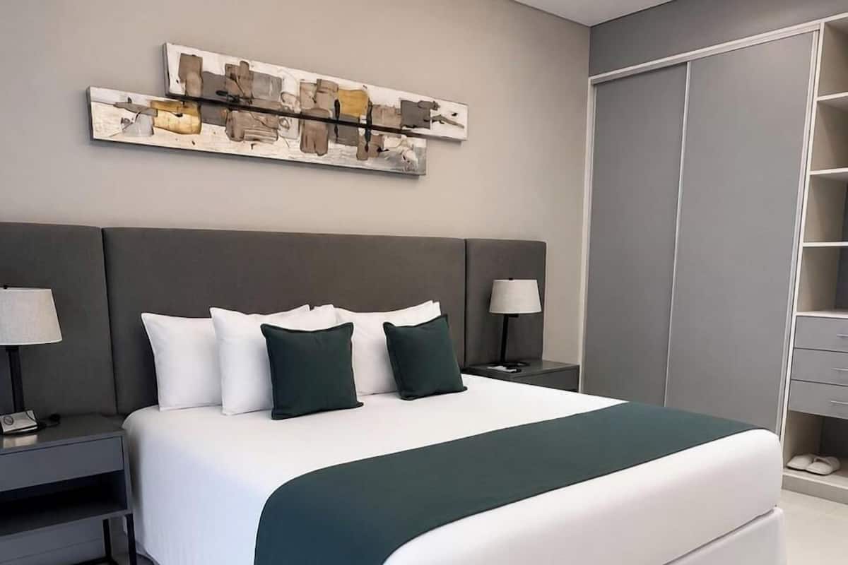 A modern hotel room with a large bed, grey headboard, two bedside tables with lamps, a grey wardrobe, and a wall art piece.