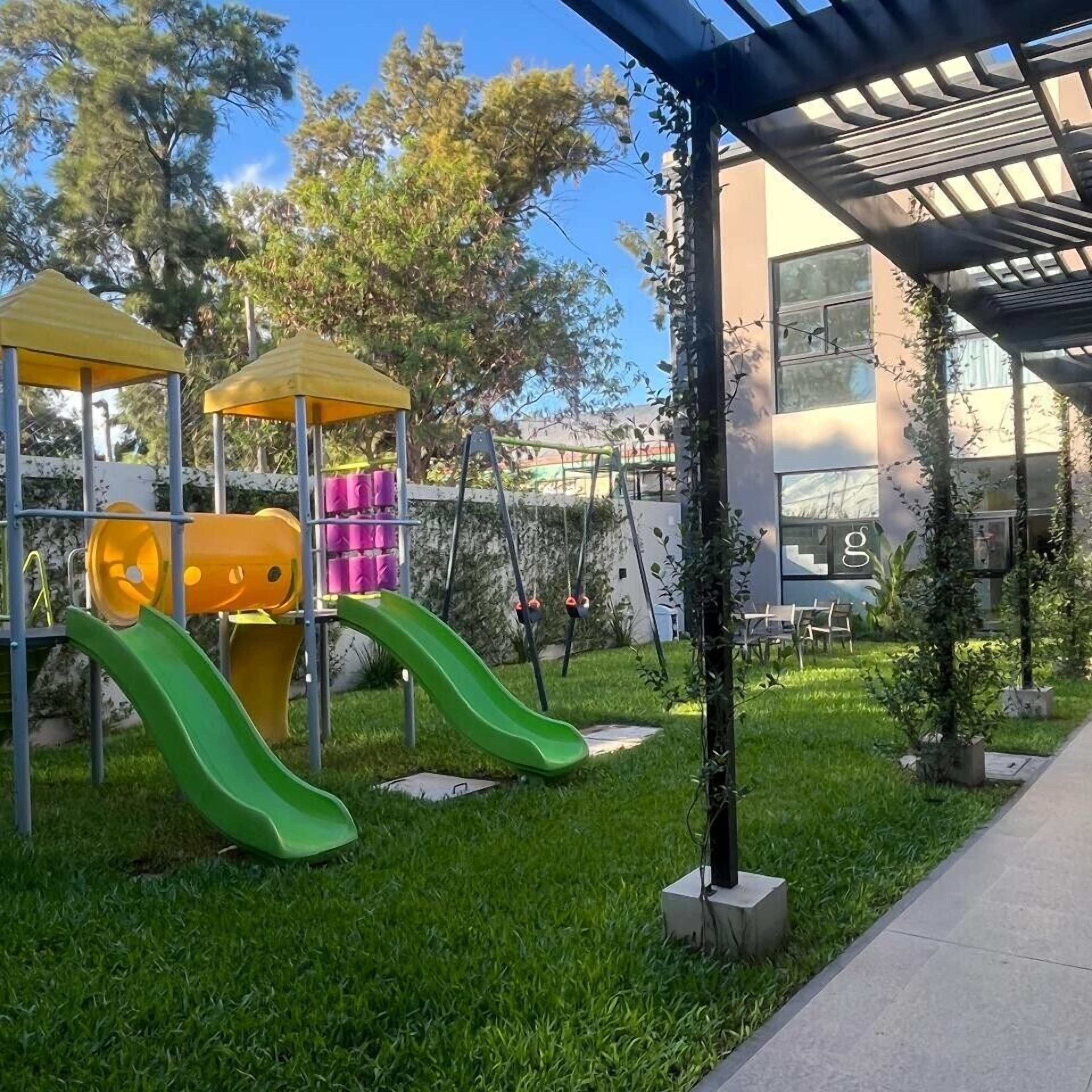 A playground with slides and climbing structures, surrounded by grass and trees.