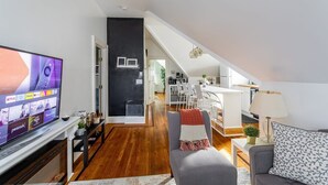 A modern living room with a flat-screen TV, a grey sofa, and a dining area with white furniture. - Renovated 2BR Retreat / Modern Comforts Included (Bridgeport)