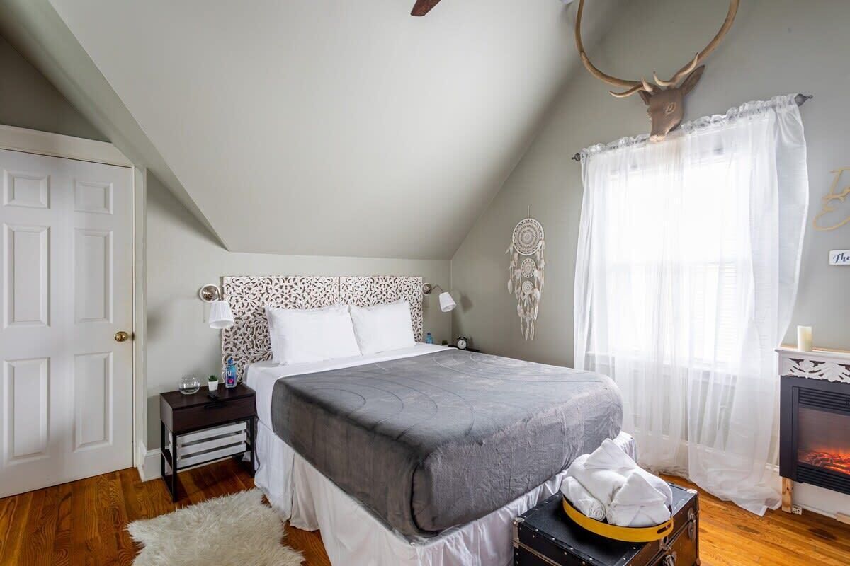 A bedroom with a bed, a nightstand, a dresser, a fireplace, and a deer head on the wall.