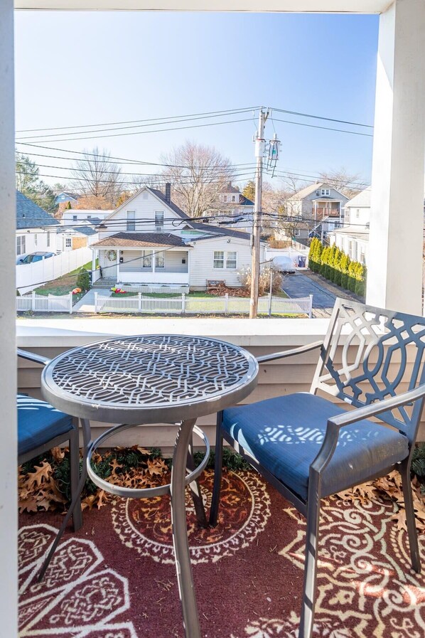 A balcony with a round table and two chairs, a patterned rug, and a view of a residential area with houses and a white picket fence. - Sunny & Spacious 2BD, 1BA WFH Apt-Huge Yard & Patio (Fairfield)
