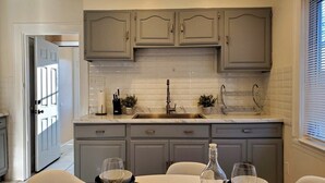 Una cocina con gabinetes grises, un salpicadero blanco de azulejos, un fregadero y un comedor con mesa y sillas.