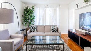 Una sala de estar con un sofá, sillones, una mesa de centro de vidrio, un televisor y una planta grande.