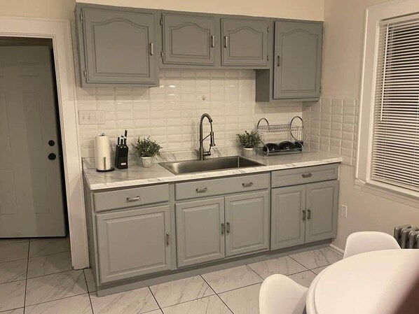 A kitchen with grey cabinets, a white tiled backsplash, a sink, and a window with blinds.