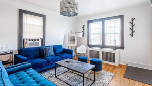 A modern living room with a blue sofa, a coffee table, and a large window with blinds. - The Blue Sapphire: Modern & Stylish 2BR w/ 2 King Beds & Full Kitchen (Bridgeport)