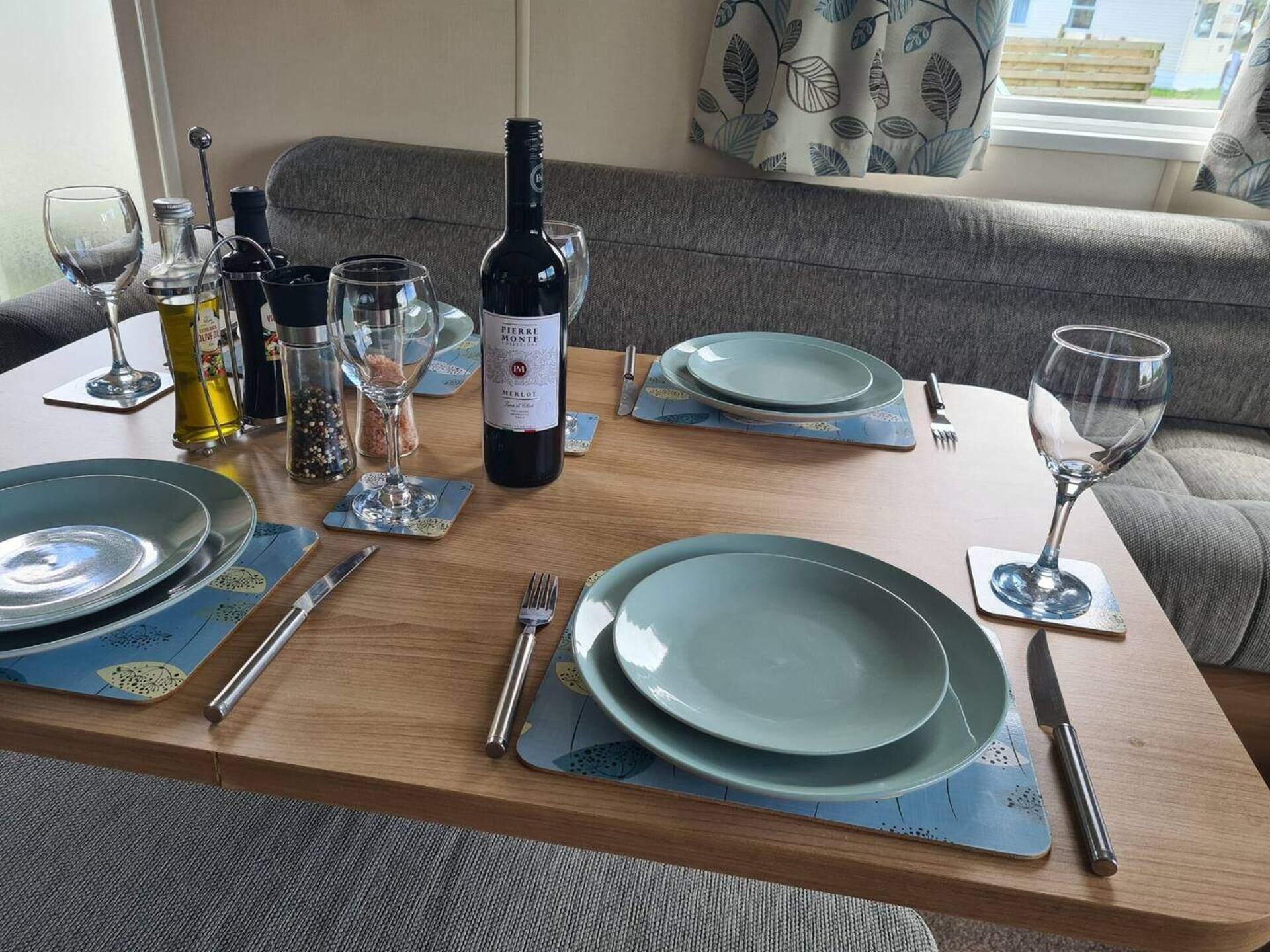A dining table set with plates, cutlery, glasses, and a bottle of wine, with a sofa and curtains in the background.