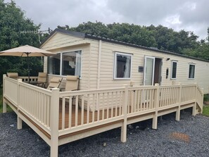 Une maison mobile dotée d’une terrasse, d’un espace extérieur couvert avec des sièges et d’une fenêtre sur laquelle est affiché un panneau indiquant « MOONS ».
