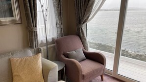 A living room with a sofa, armchair, and a view of the sea through large windows. - Lavender super front row sea view caravan at Rockley Park, Poole Harbour, Dorset (Poole)