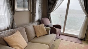 A living room with a sofa and armchairs, a large window with curtains, and a view of the sea. - Lavender super front row sea view caravan at Rockley Park, Poole Harbour, Dorset (Poole)