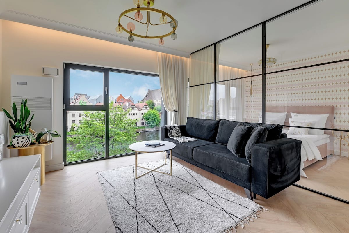 A modern living room with a black sofa, a round coffee table, and a view of buildings through large windows.