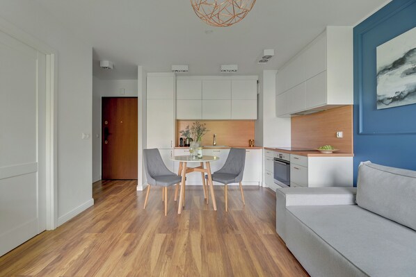 Una cocina moderna con armarios blancos, un comedor con dos sillas grises y una mesa redonda, y un sofá gris.