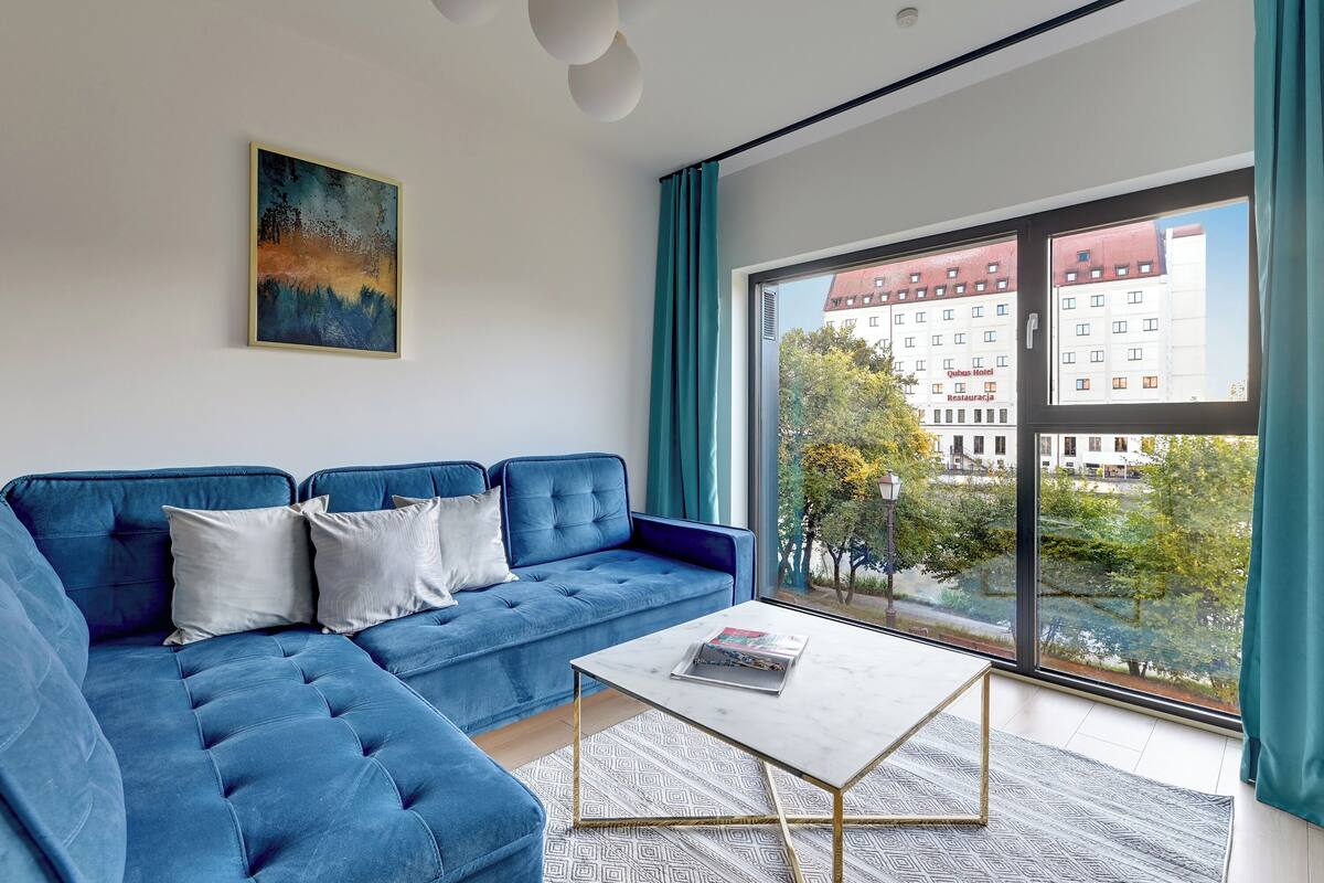 A modern living room with a blue sofa, a white coffee table, and a large window with a view of buildings and trees.