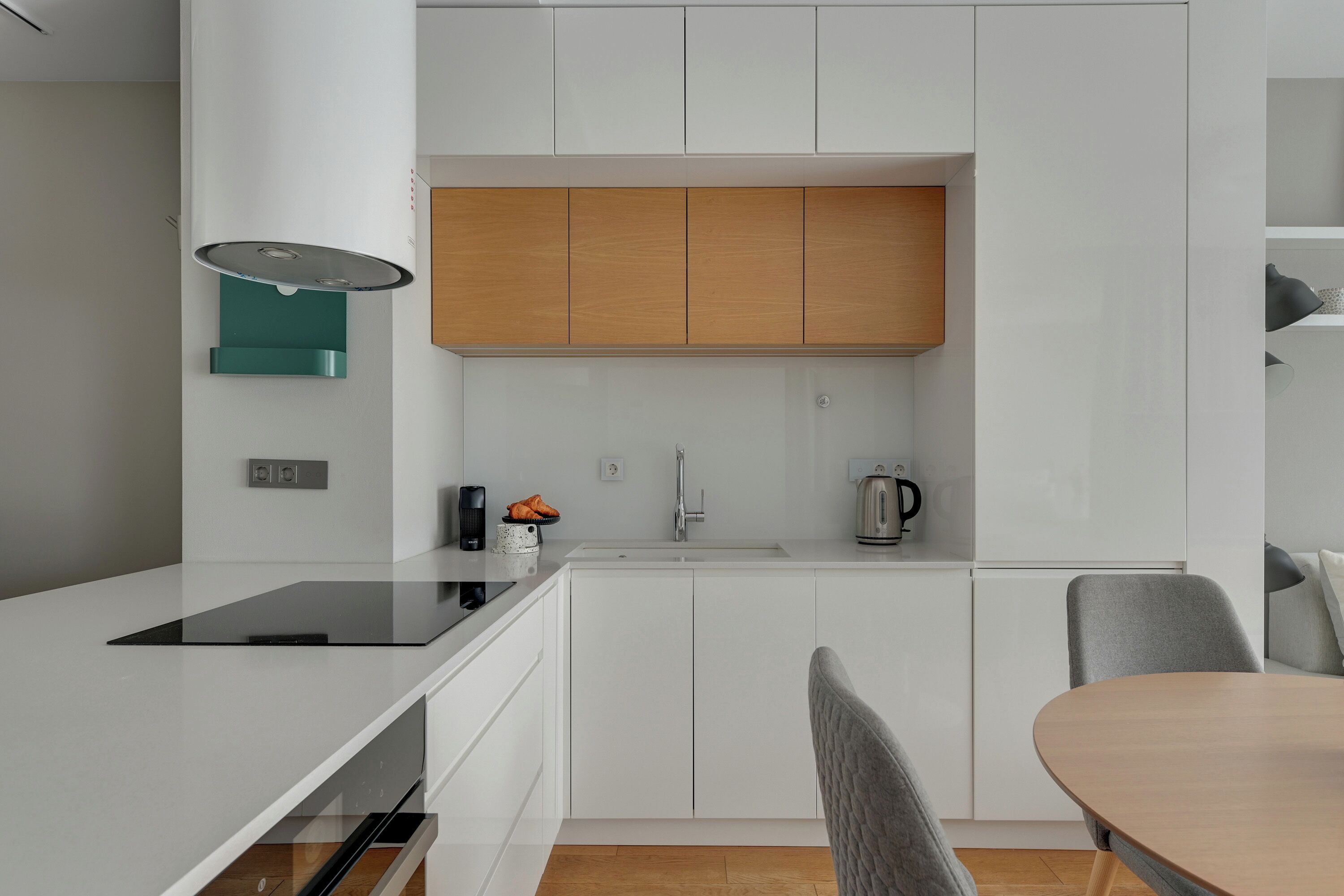 A modern kitchen with white cabinetry, a wooden upper shelf, a built-in stove, and a dining area with a table and chairs.