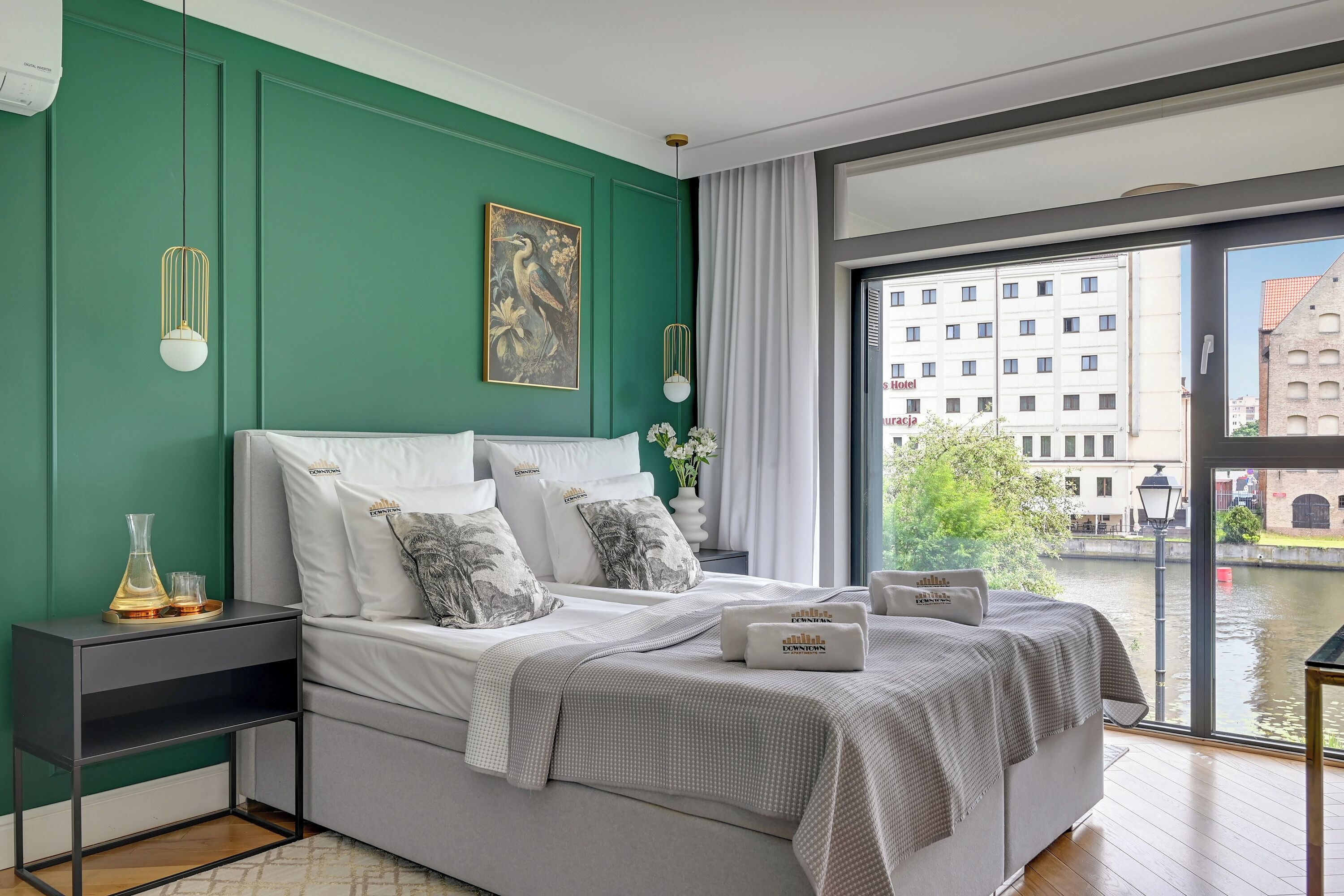Ein modernes Schlafzimmer mit grüner Wand, einem großen Bett mit weißer Bettwäsche, einem Stadtbildgemälde und Blick auf ein Gebäude durch ein Fenster.