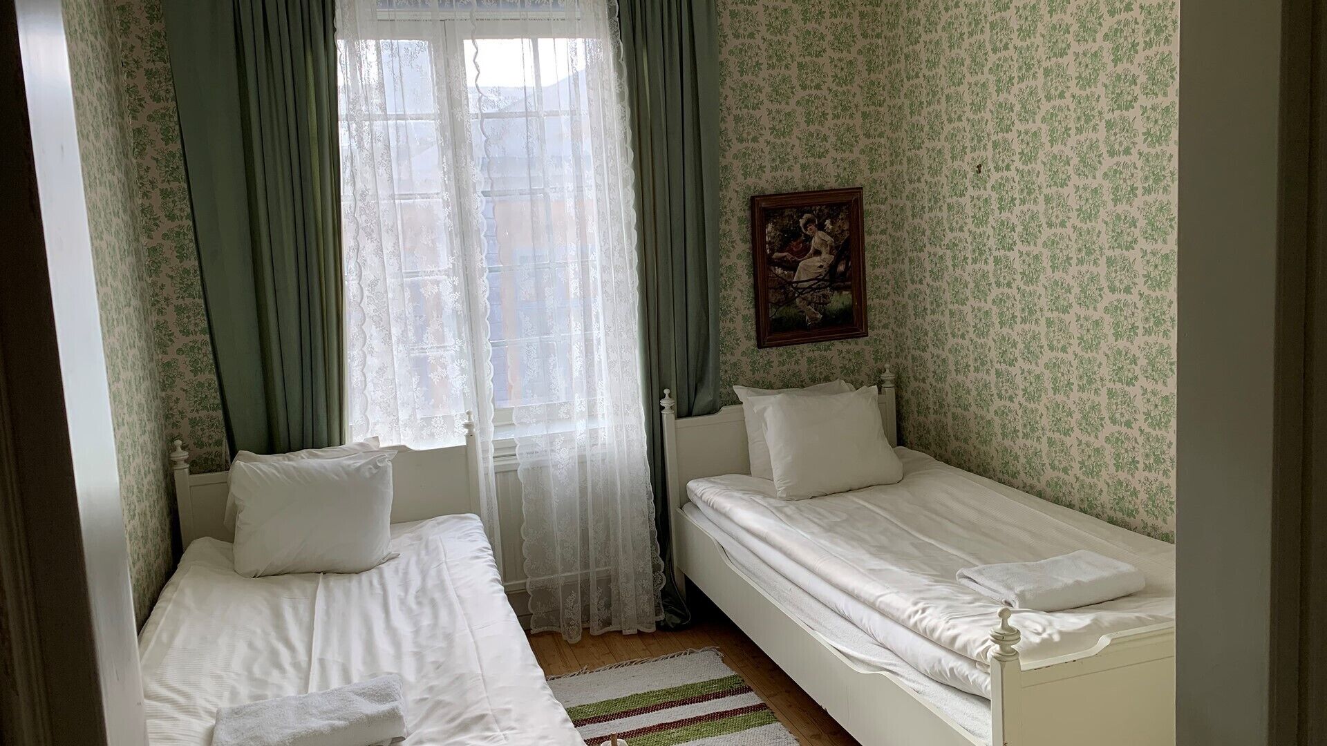 Ein Zimmer mit zwei Einzelbetten, einem Fenster mit leichtem Vorhang und blumigem Wandpapier.