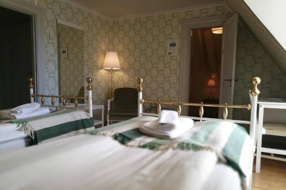 Une chambre d’hôtel avec deux lits, une couvre-lit à rayures vertes et blanches, une tête de lit blanche et une lampe sur une table de chevet.