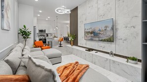 A modern living room with a large sectional sofa, a flat-screen TV mounted on a marble wall, and a kitchen in the background. - 5BD/5BA Townhouse with Pool, Theater & Games (Orlando)