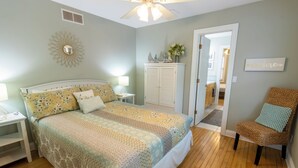 Un dormitorio con una cama, mesitas de noche, un aparador, una silla y una puerta que da a otra habitación.