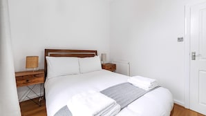 Una cama bien hecha con sábanas blancas y una toalla doblada, una mesita de noche de madera con una lámpara y una puerta blanca.