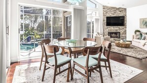 A spacious living room with a round glass table, wooden chairs, a fireplace, and a large TV.
