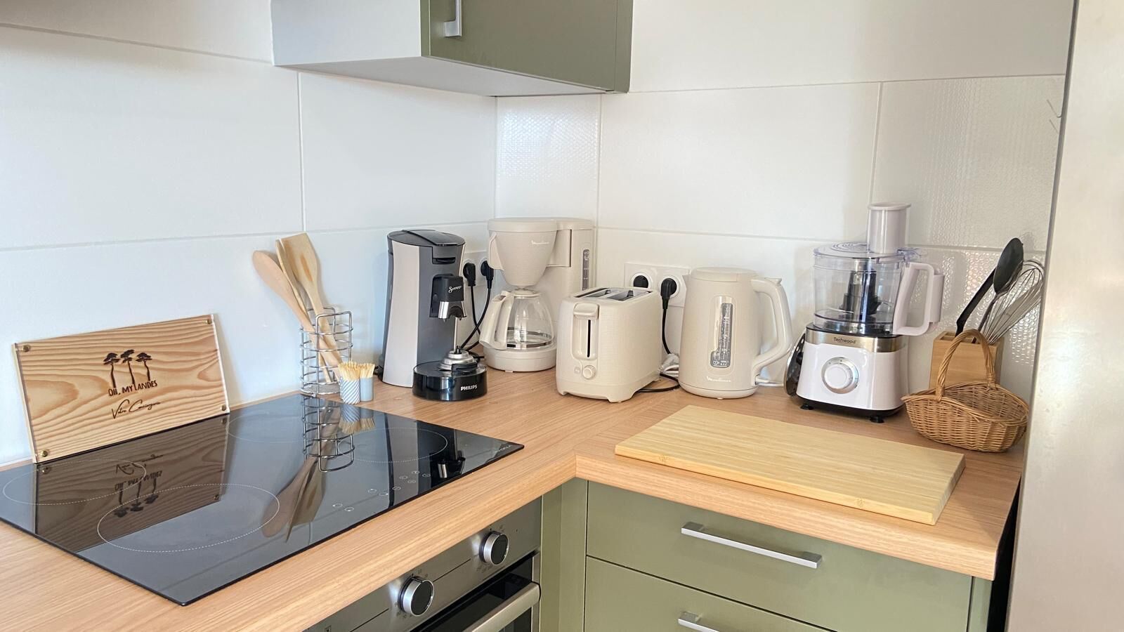 A compact kitchen with a black induction cooktop, a coffee maker, a toaster, a blender, and a wooden cutting board.