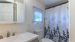 A bathroom with a white sink, a toilet, a window with blinds, and a shower curtain with a floral pattern. - Studio apartment above Horseshoe Bay beach (Southampton Parish)