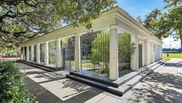 Un pavillon couvert, doté de colonnes blanches et d’une clôture en métal, entouré d’arbres et d’une pelouse.