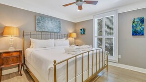 Une chambre à coucher comprenant un lit, des tables de chevet, un ventilateur de plafond, des œuvres d’art et une fenêtre avec des stores.